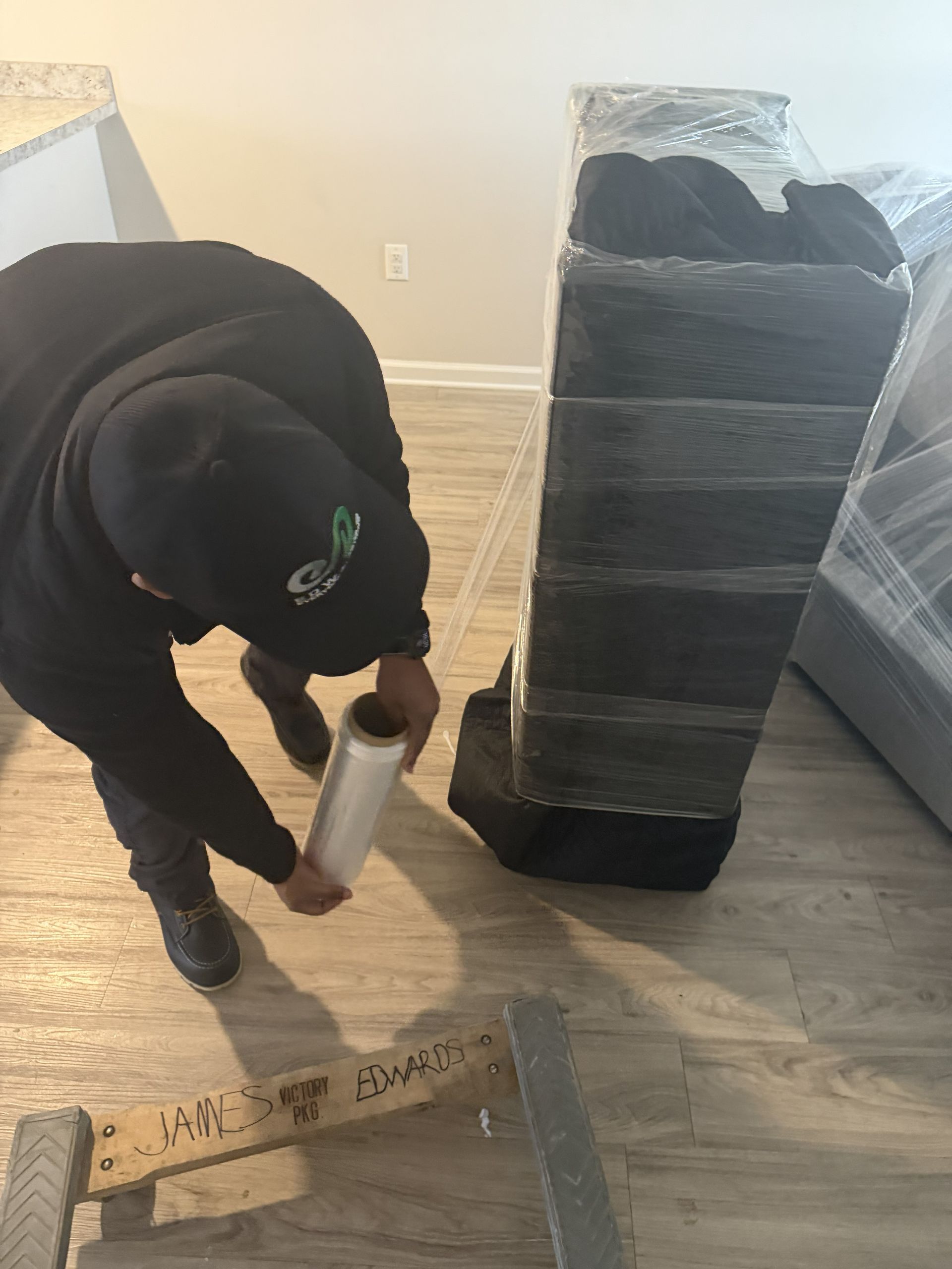 A person in a black hoodie and beanie wraps a tall, black furniture item in clear plastic film in a room with wood floors.