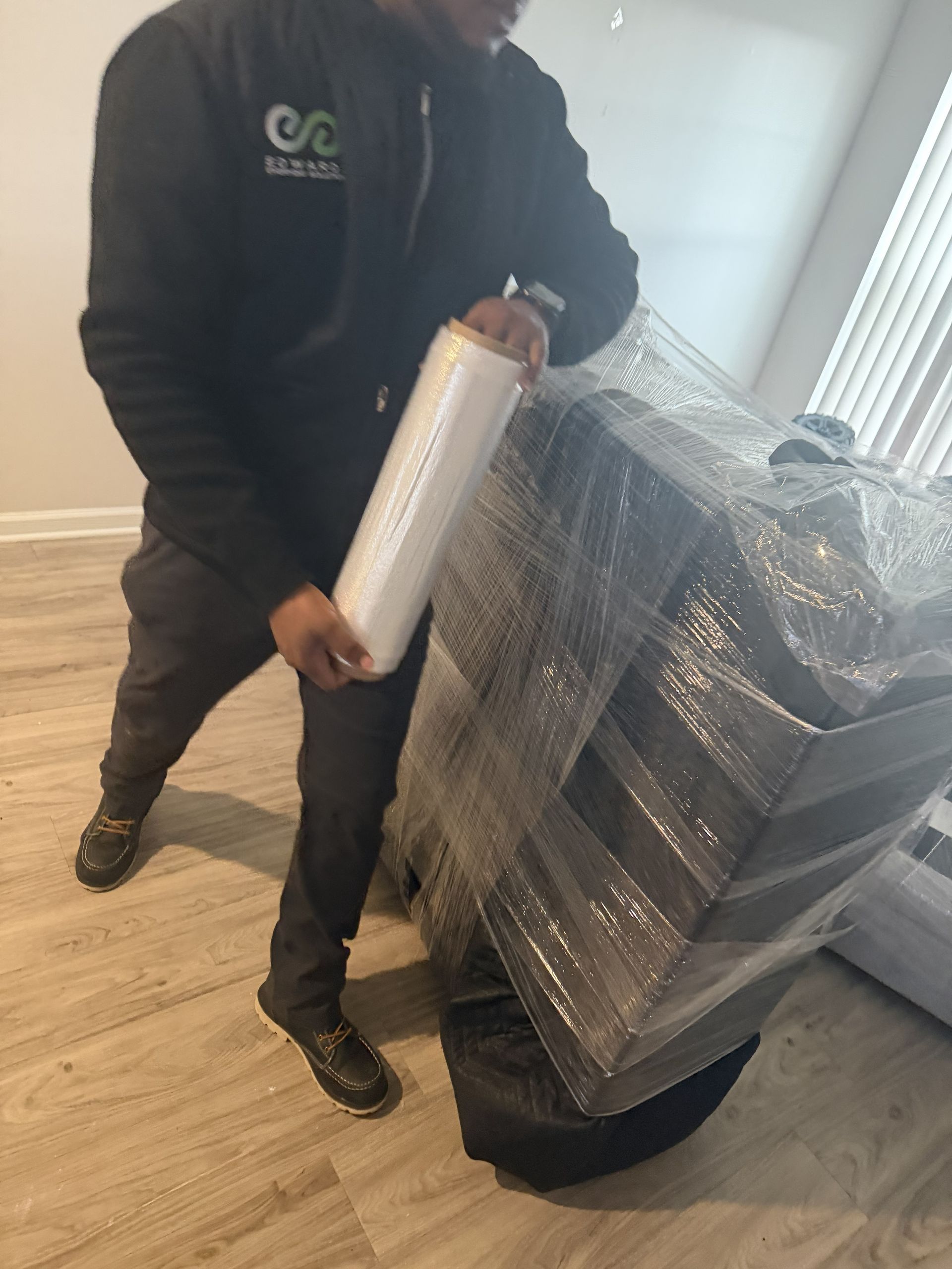 A person in a black jacket holds a roll of plastic stretch wrap next to a piece of furniture wrapped in bubble wrap.