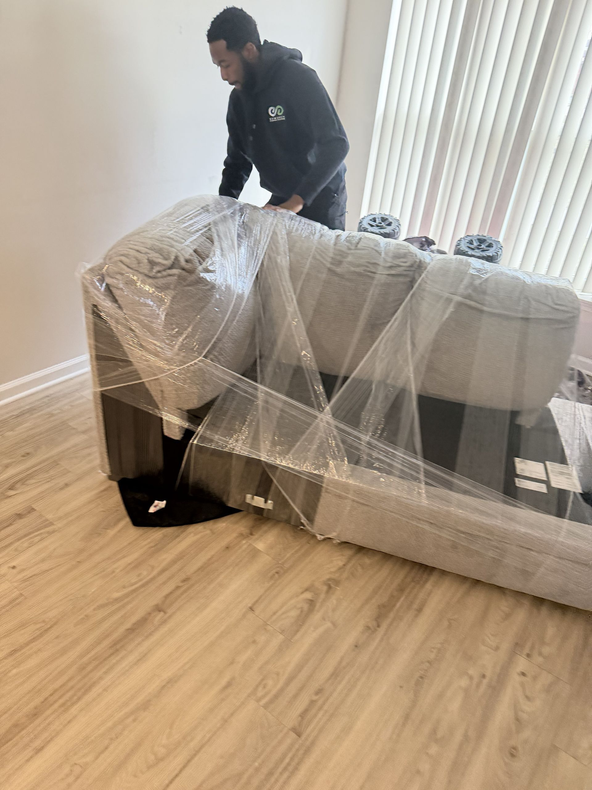 A person wraps a light-gray sofa in clear plastic film inside a room with light wood flooring and vertical blinds.