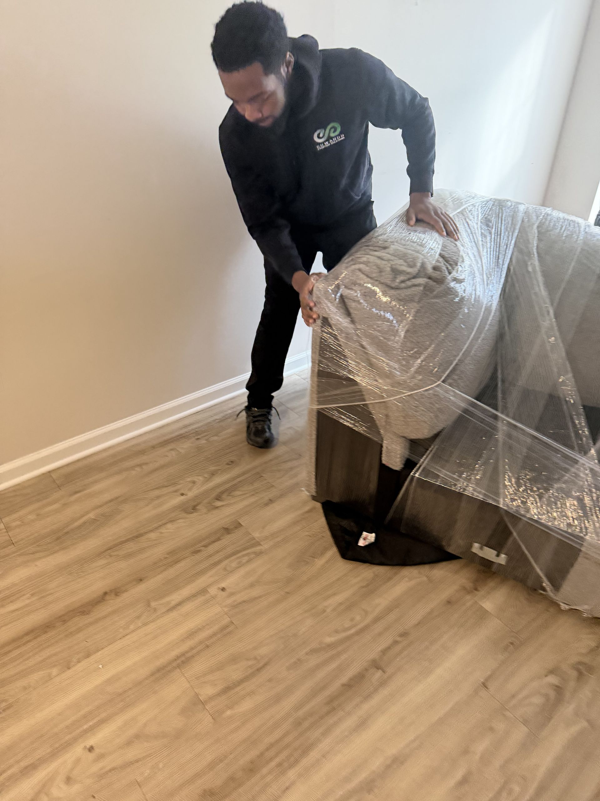A person in dark clothing unwrapping a piece of furniture covered in bubble wrap inside a room with wood-look flooring.