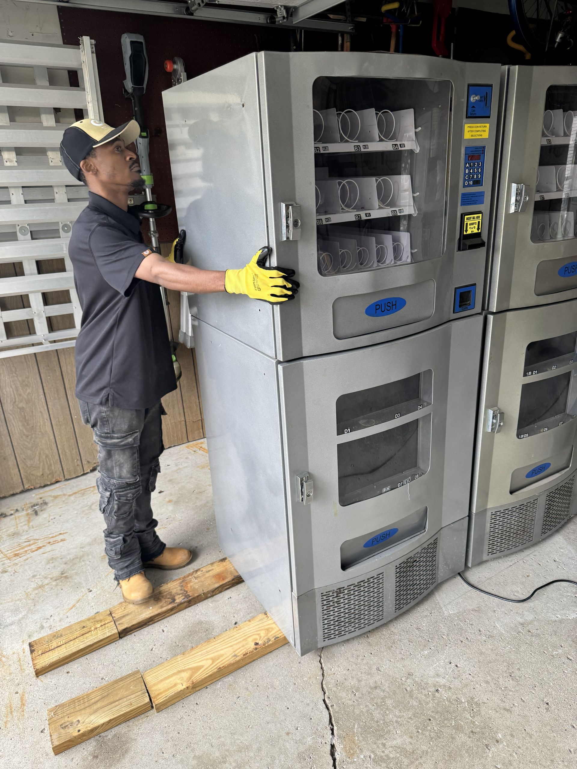 A person in work clothes and yellow gloves guides a large gray vending machine resting on wooden planks in a garage.