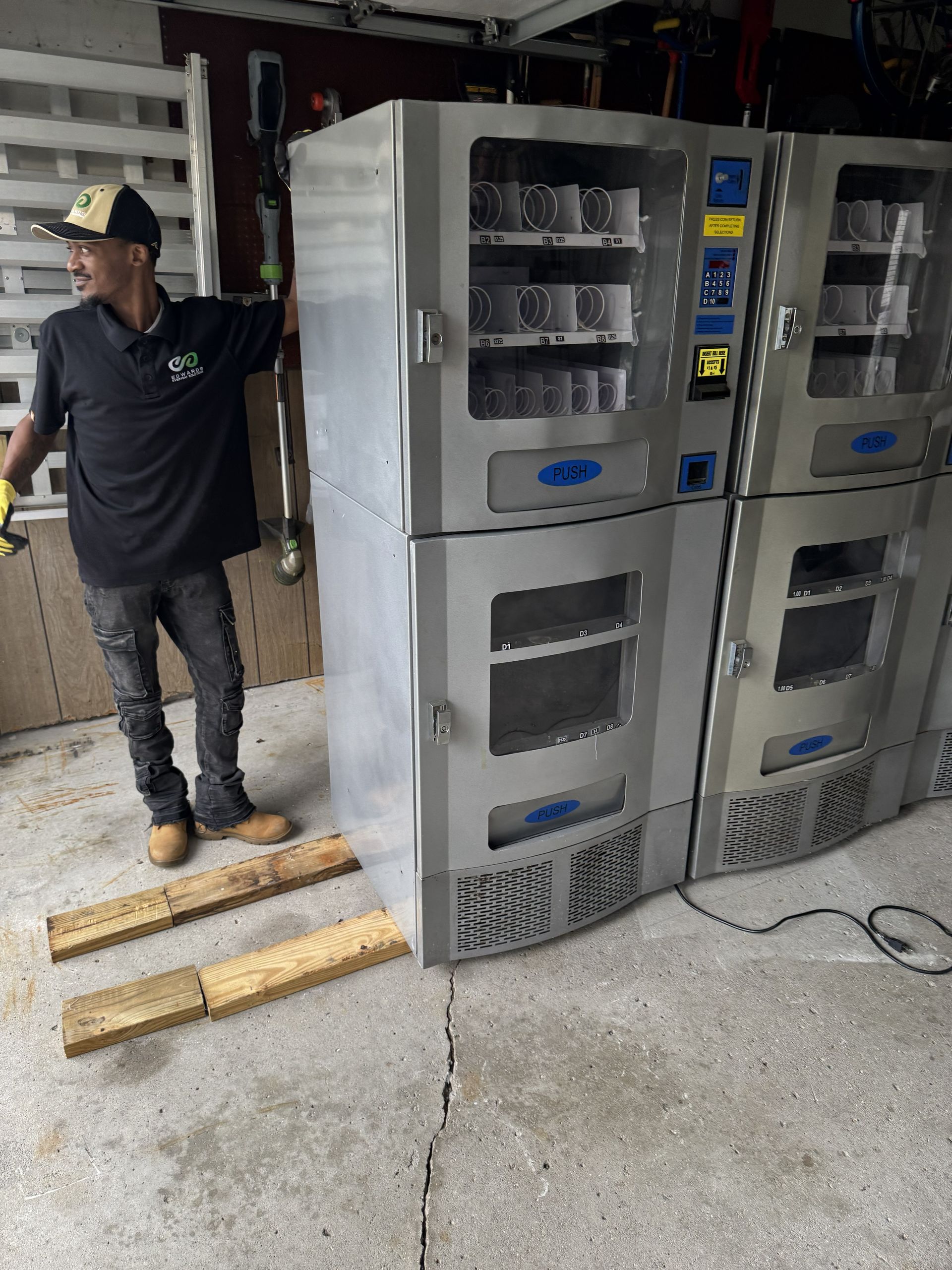 A person stands beside two stacked stainless steel vending machines placed on wooden blocks in a storage area.