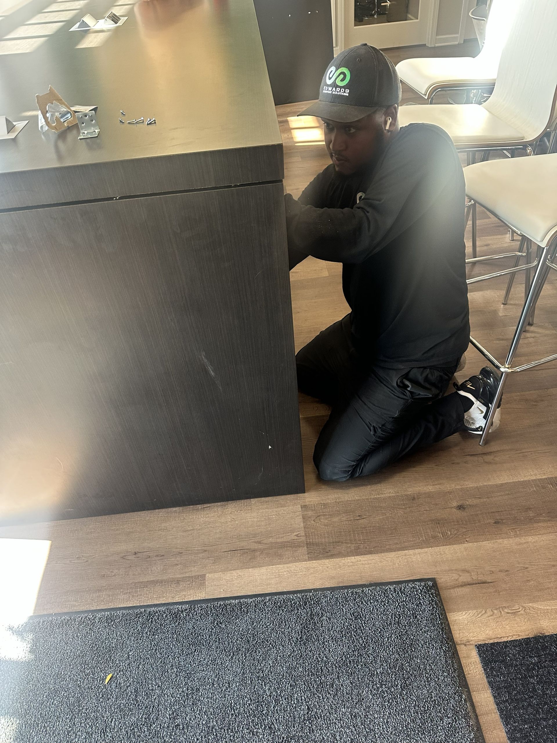 A person in a dark uniform and cap kneels on a wooden floor, working on the side of a large, dark-colored counter.