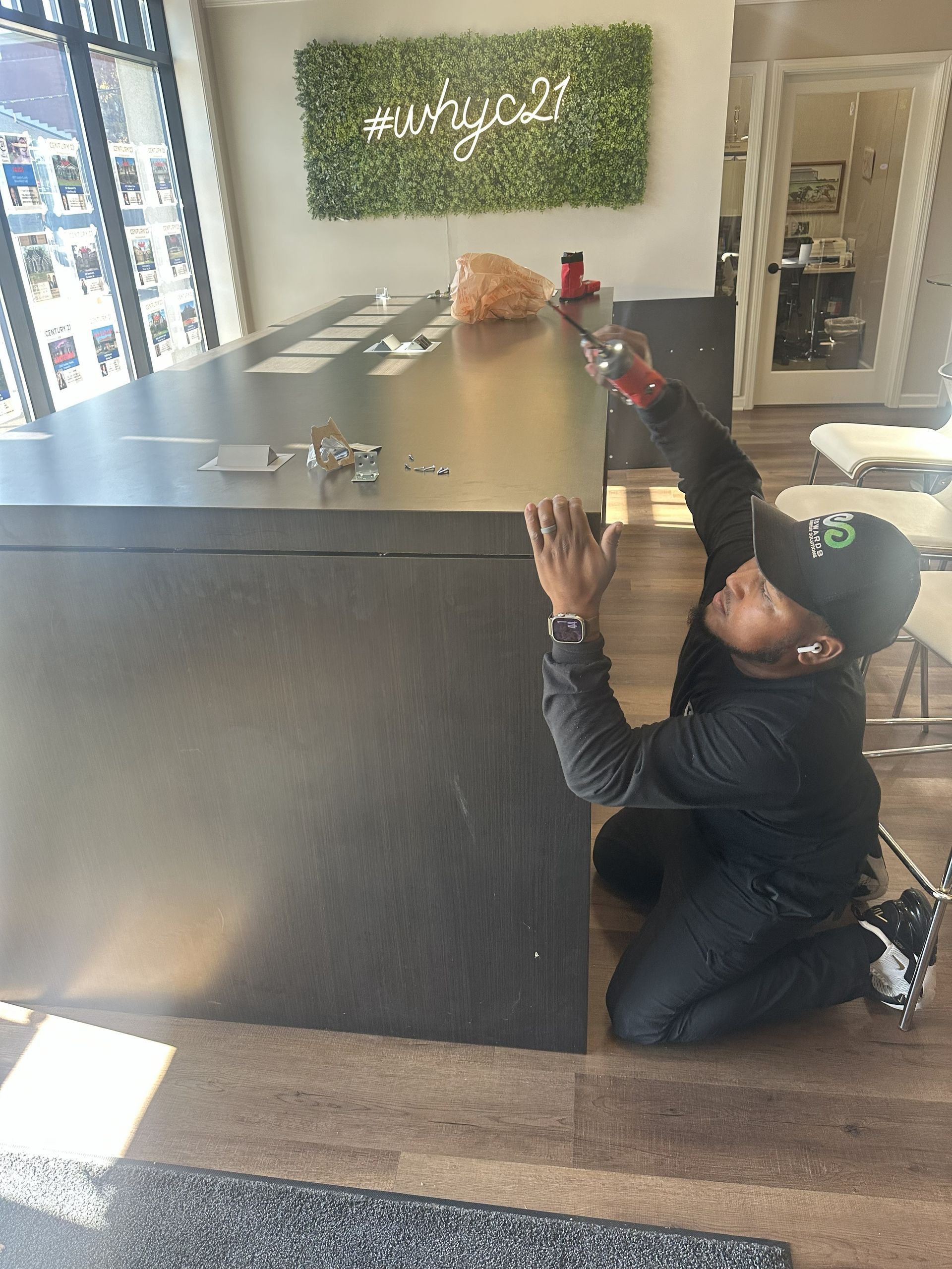 A person in a black shirt and cap kneeling by a large dark counter, spraying it with a cleaning product in a shop.