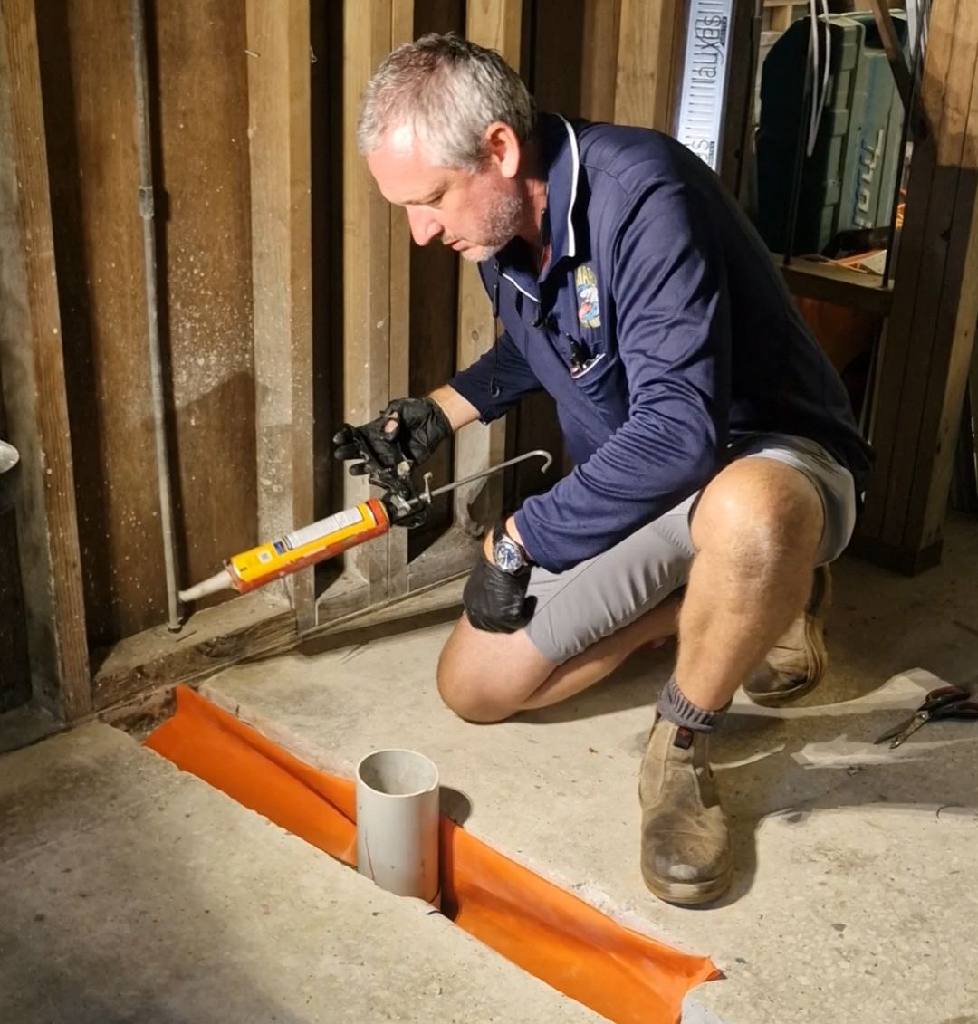 Man applying sealant near an orange drain in a basement. He kneels, using a caulking gun — Shark Pest Control in Peregian Beach, QLD