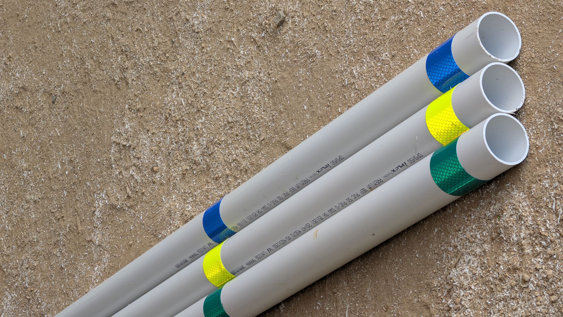 Three gray pipes lie on a sandy surface, each marked with a different color band: blue, yellow, and green.
