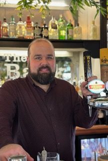 A man with a beard is standing at a bar holding a beer tap.