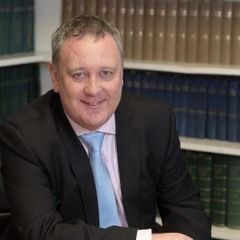 A man in a suit and tie is sitting in front of a bookshelf.