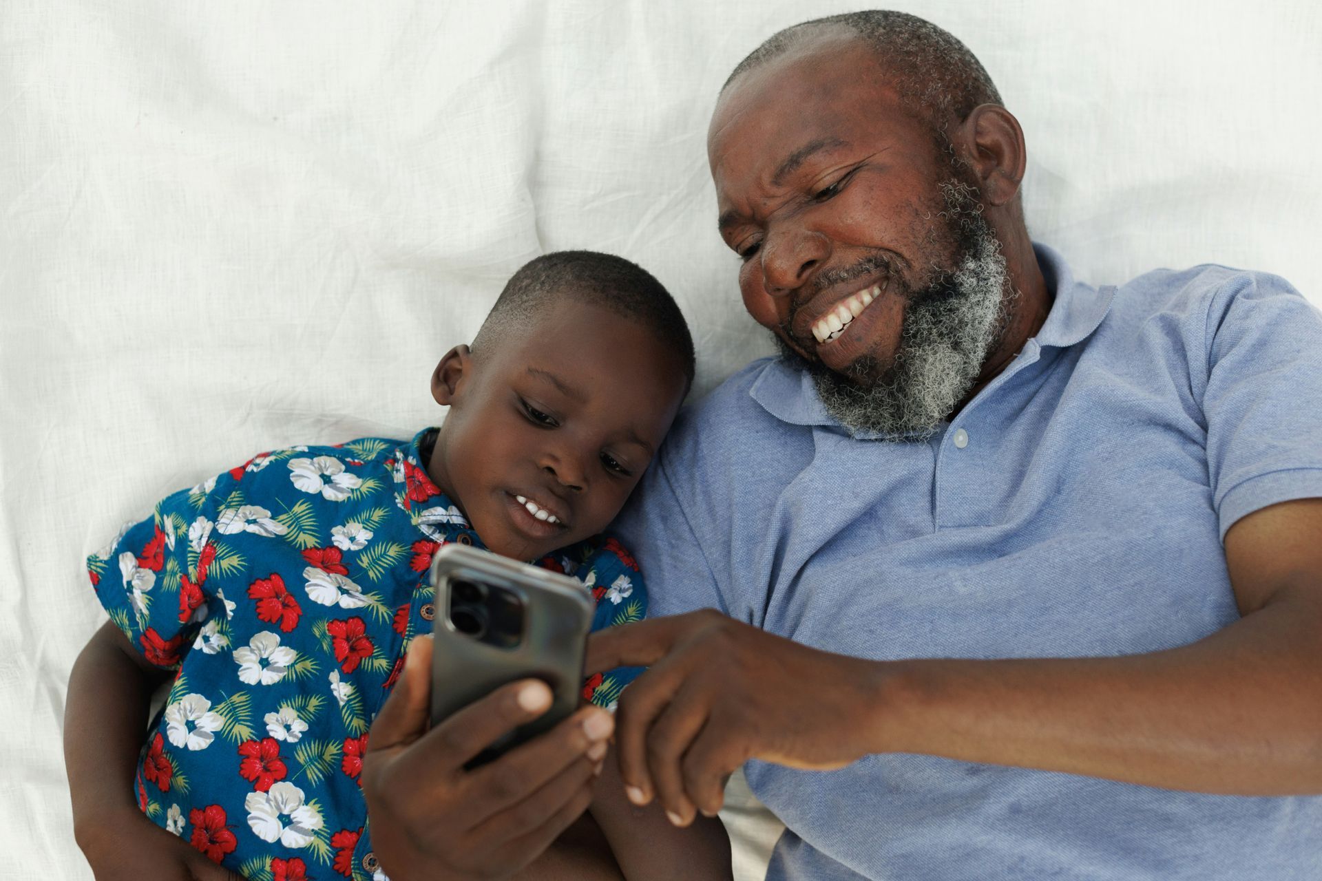 A man and a child are laying on a bed looking at a cell phone.