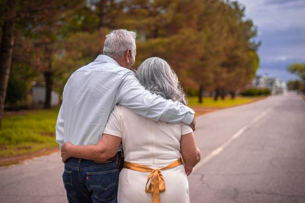 An elderly couple is walking down a road with their arms around each other.