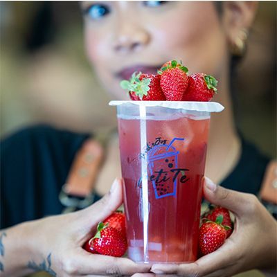 Donna che tiene in mano una tazza trasparente contenente una bevanda rossa con delle fragole in cima, e altre fragole intorno alla tazza.