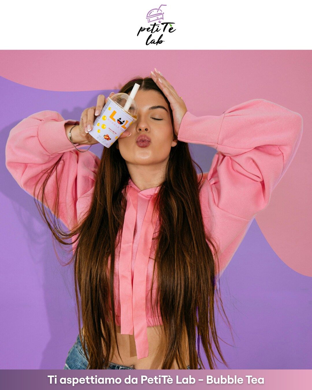 Donna con felpa rosa e cappuccio che tiene in mano un bubble tea, con le labbra arricciate, davanti a uno sfondo rosa e viola.