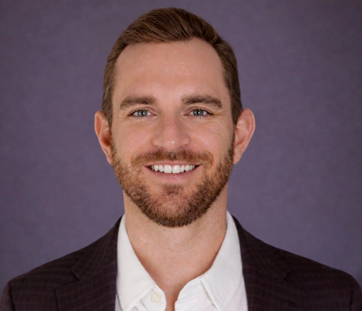A woman with long hair is smiling for the camera in front of a gray background.