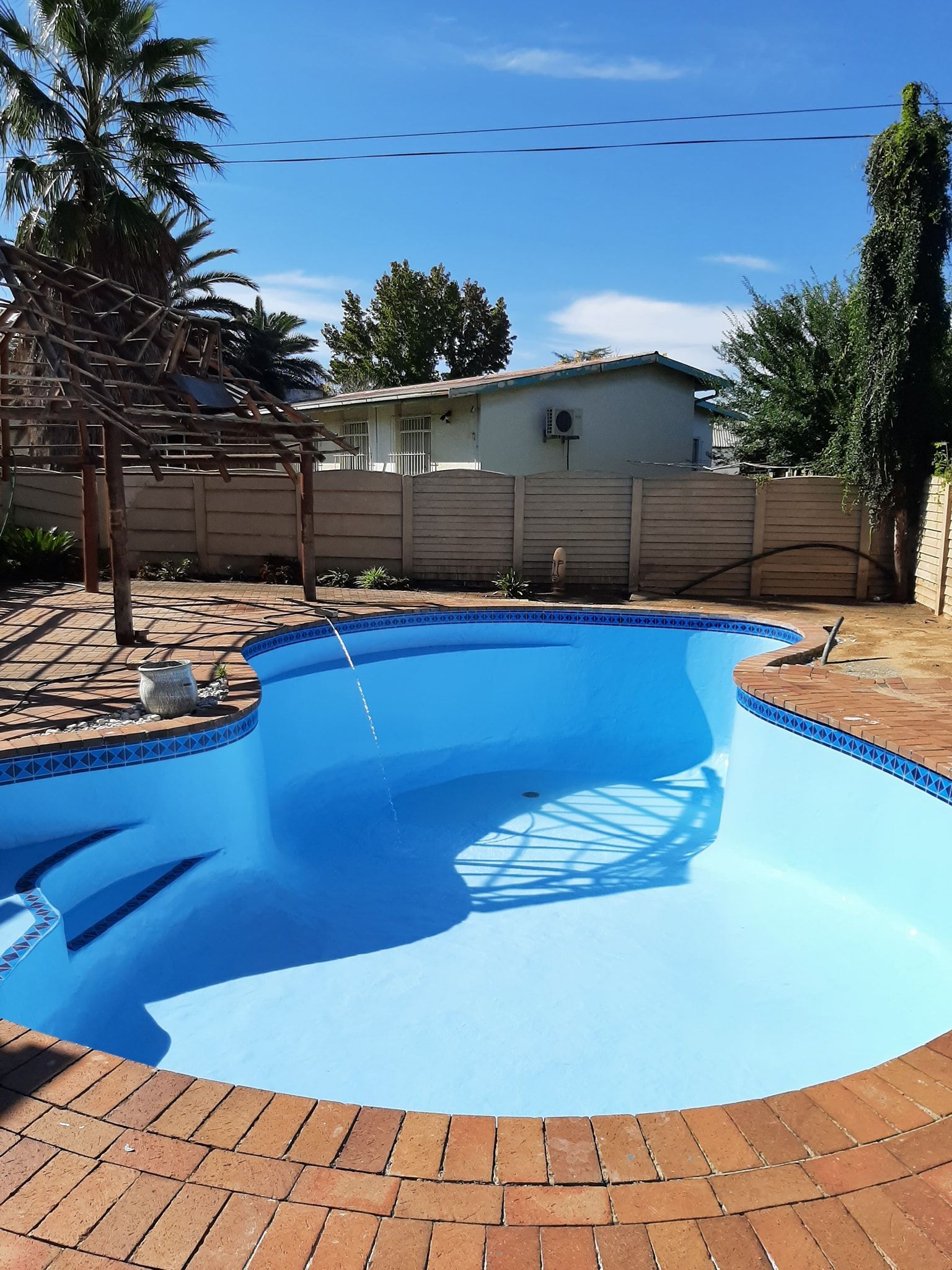 Backyard pool with bright blue water, brick patio, and wooden fence under a clear sky