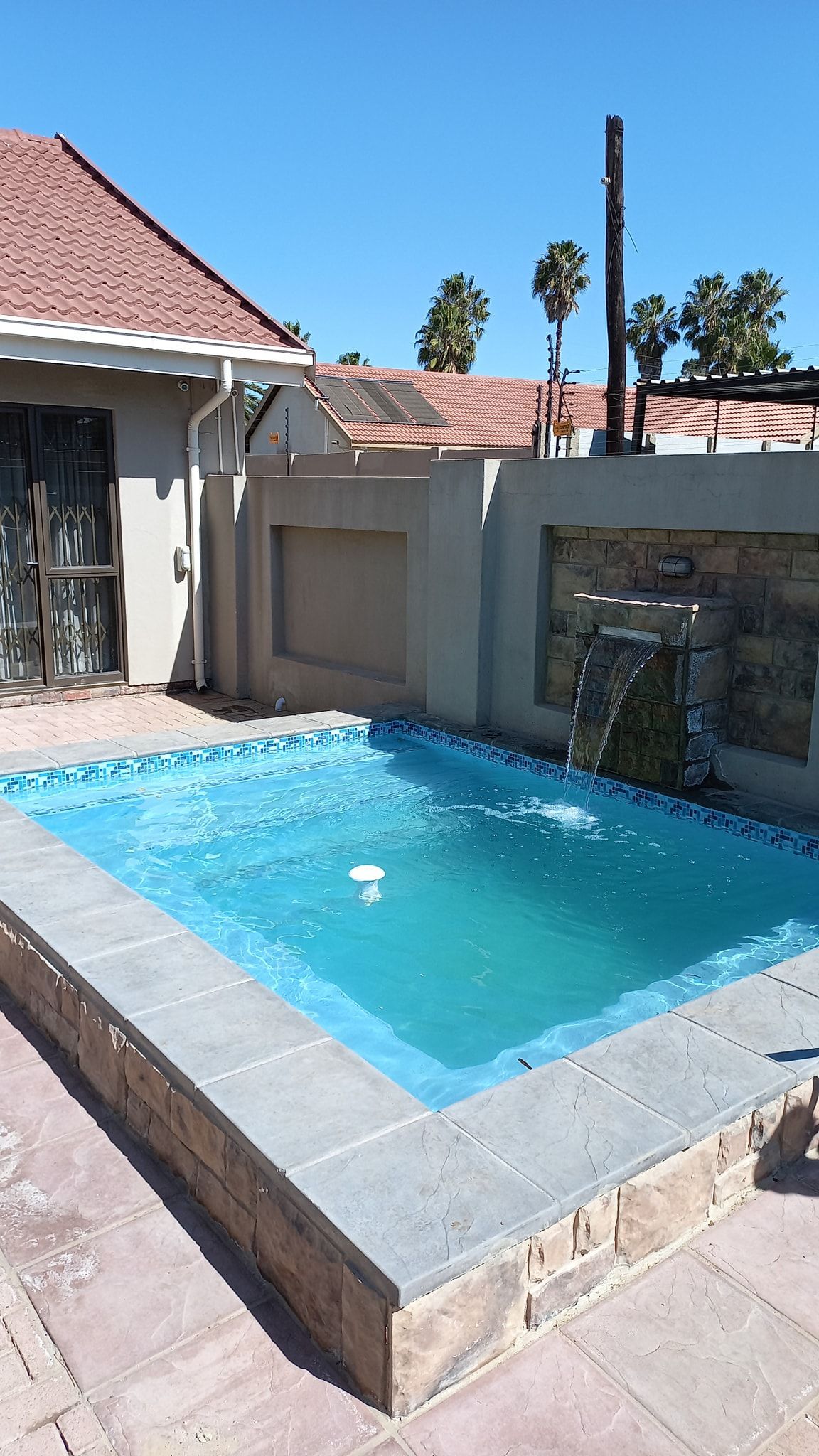 Small backyard pool with blue water beside a house, stone coping, and a tiled patio under a sunny sky