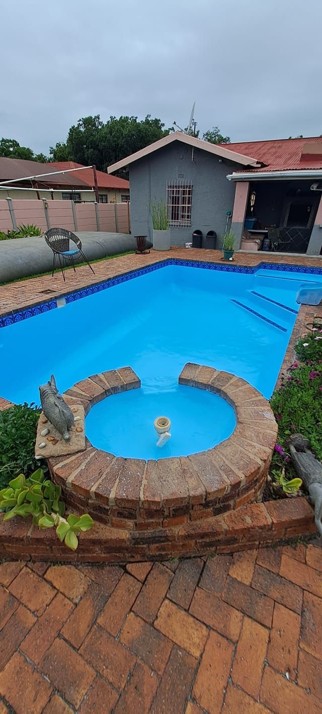 A backyard swimming pool with a circular brick-walled fountain feature in the foreground and a gray house in the back.