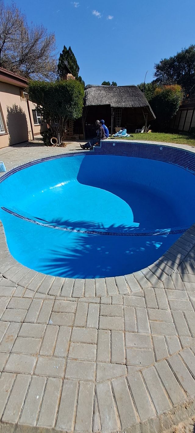 An empty, bright blue swimming pool sits surrounded by stone pavers in a sunny backyard setting.