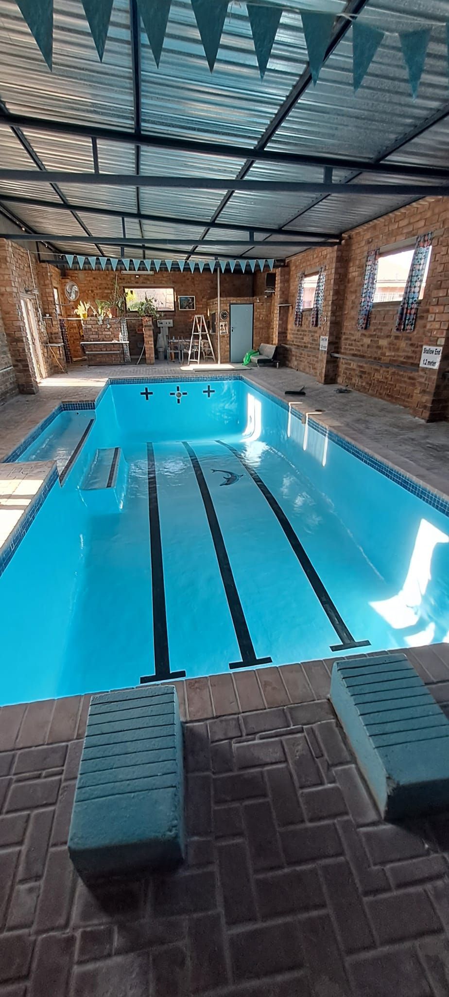 An indoor swimming pool with black lane markers, stone walls, a partially covered roof, and two foam exercise blocks.