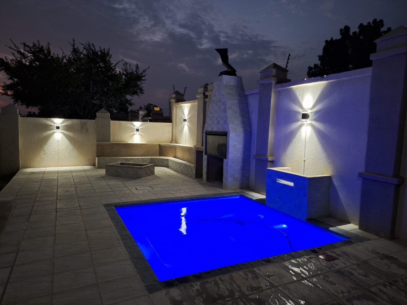 Nighttime patio with a glowing blue pool, stone deck, and wall lights behind a white perimeter wall