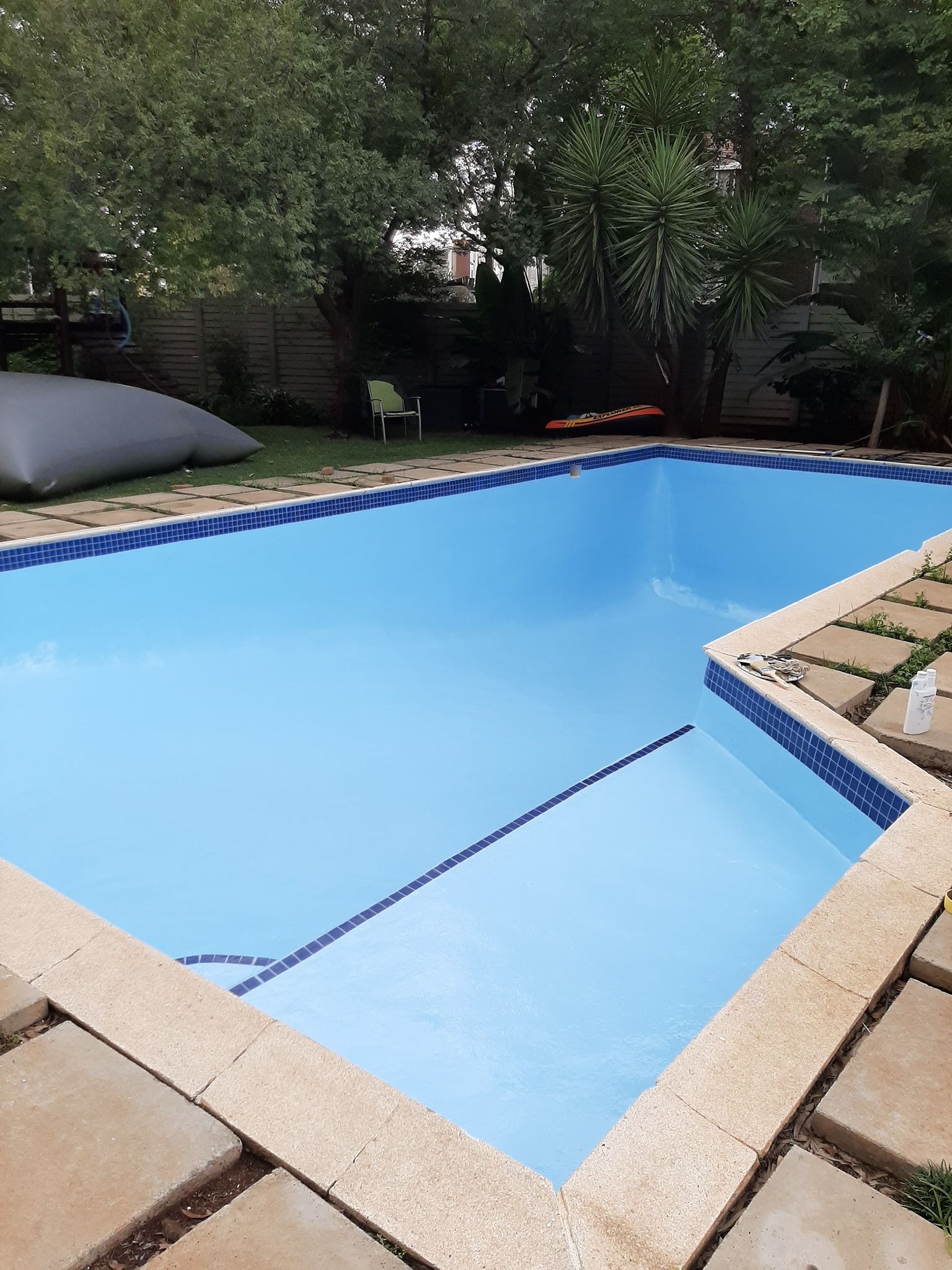 Backyard pool with bright blue cover, stone deck, and trees around it
