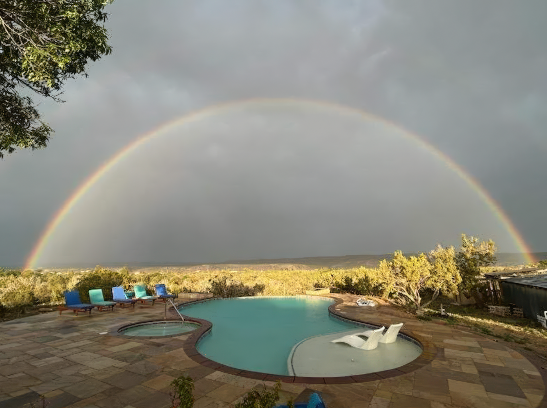 Pool and patio under a rainbow at sunset, with a cloudy sky and lounge chairs.