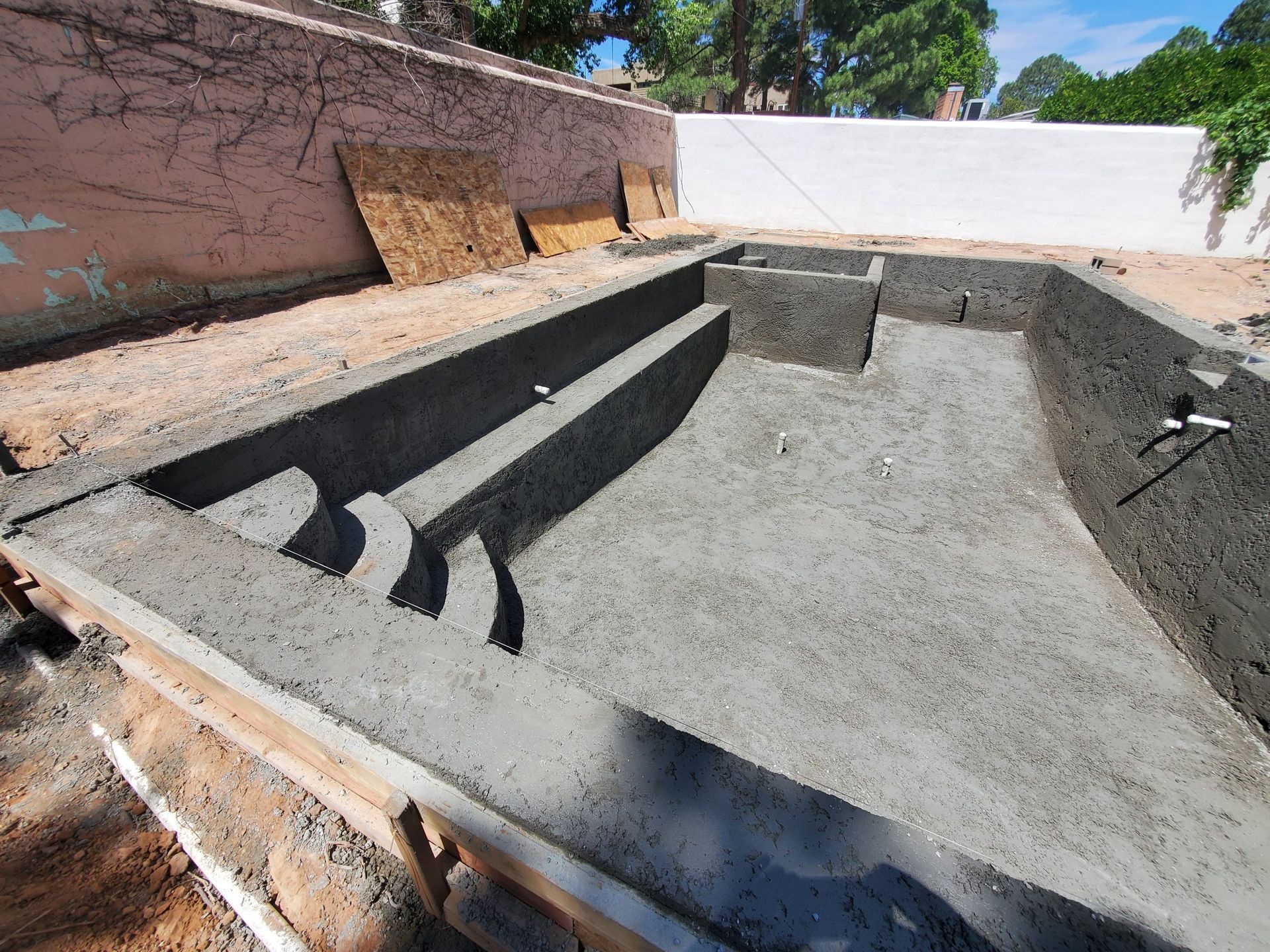 An unfinished concrete swimming pool in a backyard, featuring built-in stairs, a bench, and plumbing pipes.