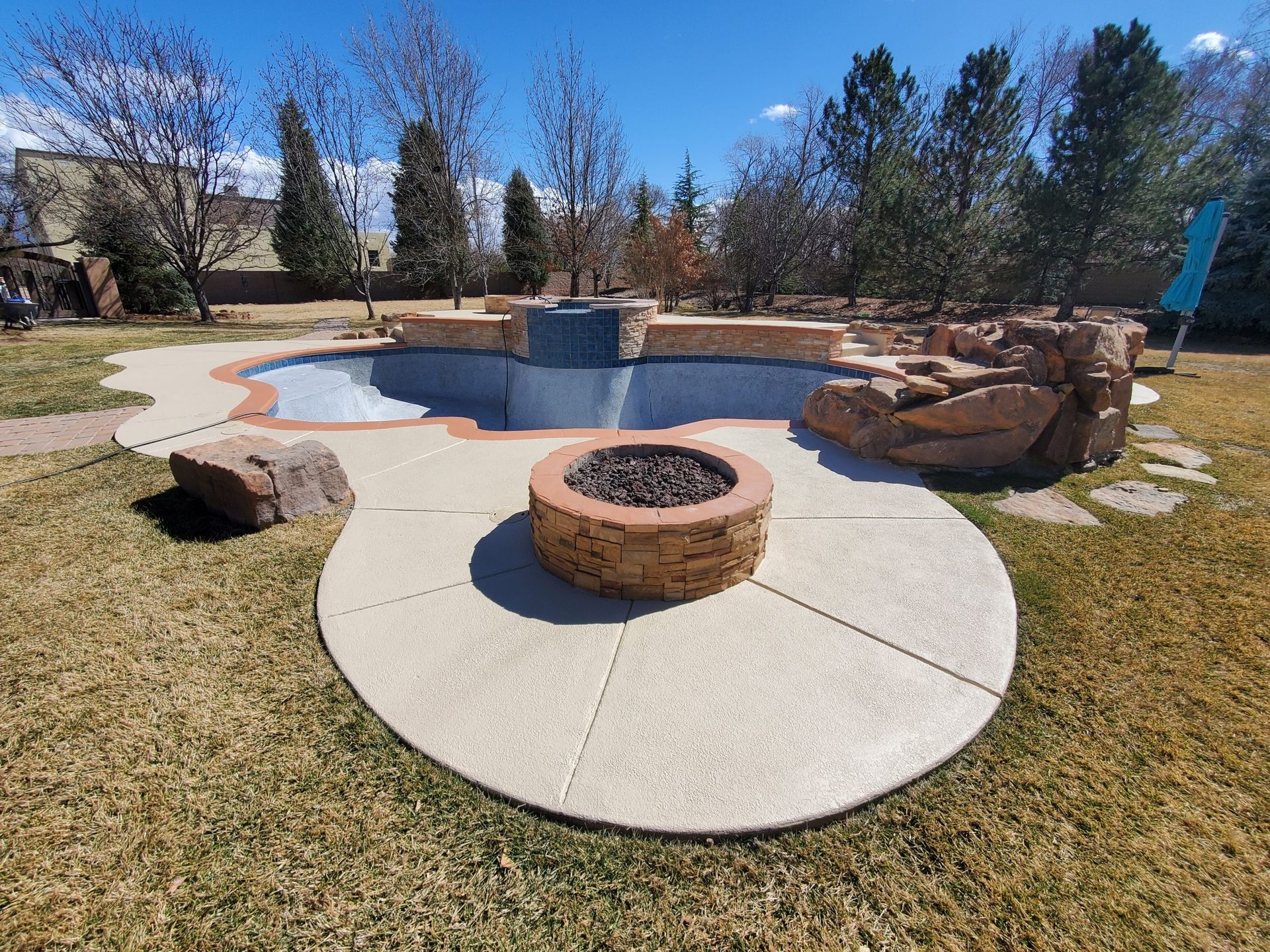 Backyard pool with stone fire pit, curving concrete patio, and landscaped trees under a blue sky