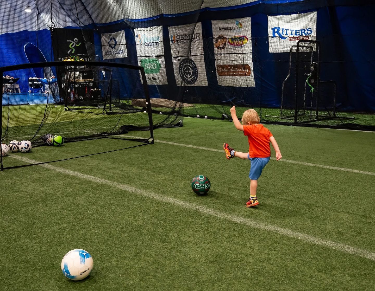 Young child kicking a soccer ball toward a net on an indoor turf field.