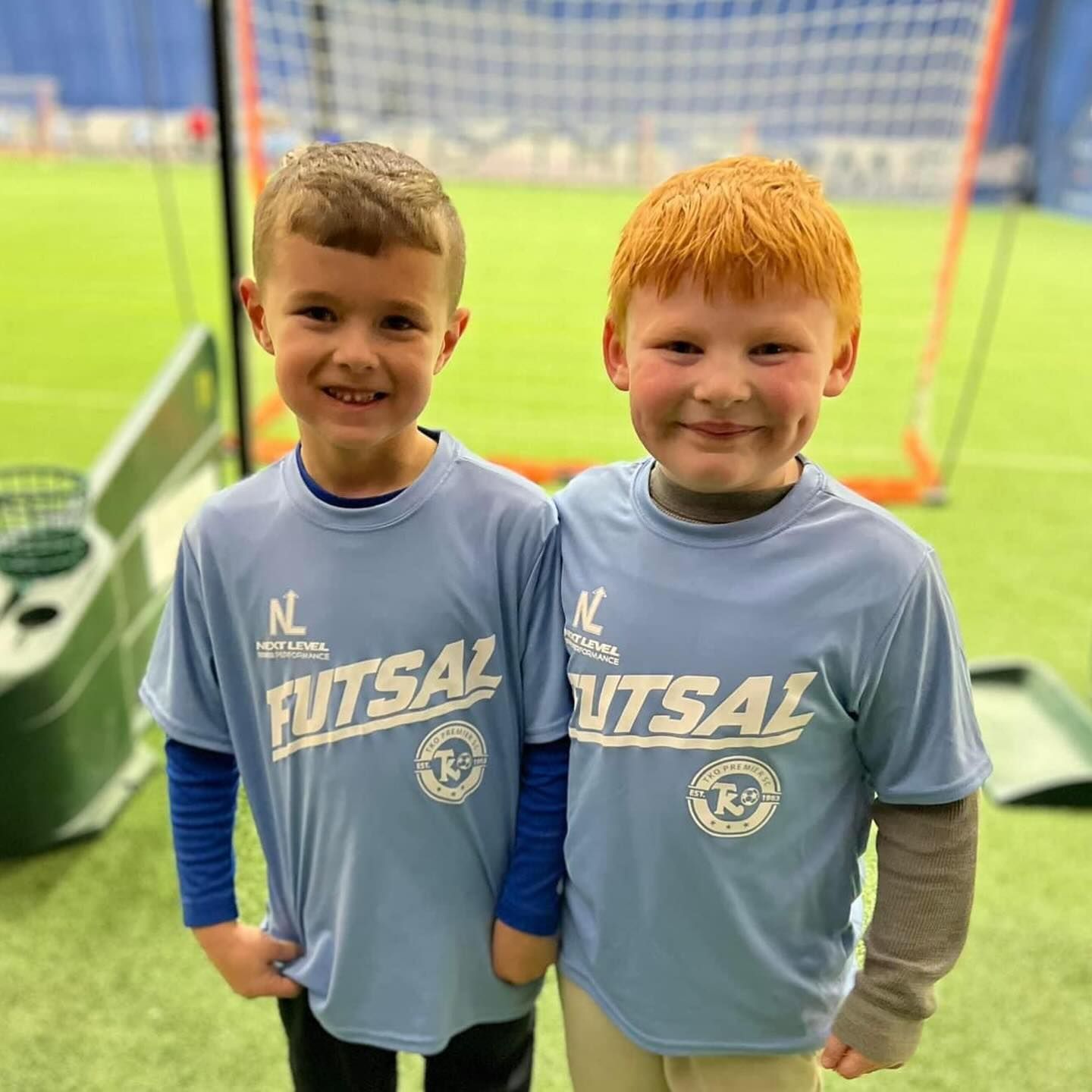 Children playing soccer indoors, wearing colored pinnies. One has a Santa hat. Green field, white and blue walls.