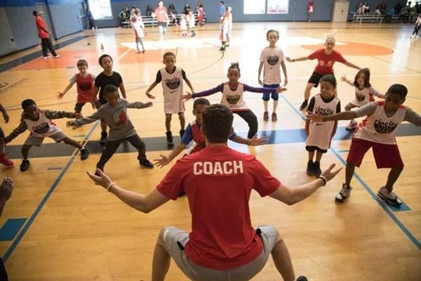 Coach leading children in exercise on a basketball court. Children are stretching with arms out. Coach in red shirt.