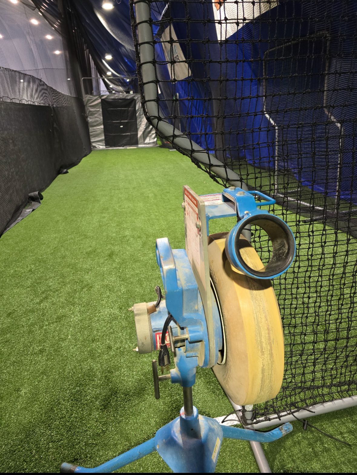 A person swinging a bat at a yellow softball inside a batting cage.