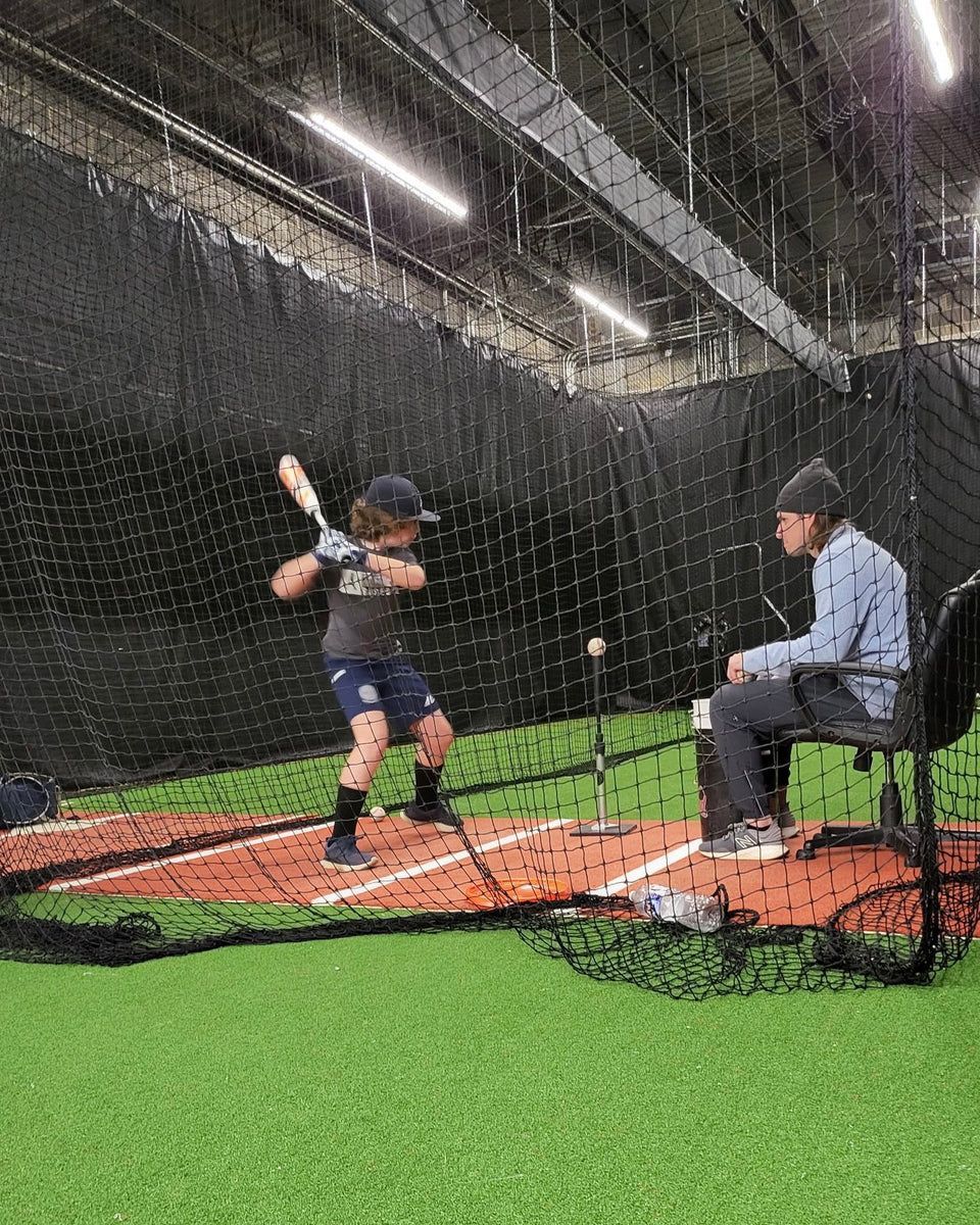 Baseball player batting at a tee, instructor watching from a chair in an indoor batting cage.