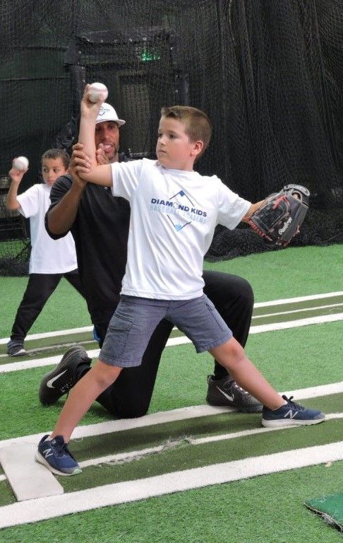A coach assisting a boy with his baseball pitching form indoors, another child in the background.