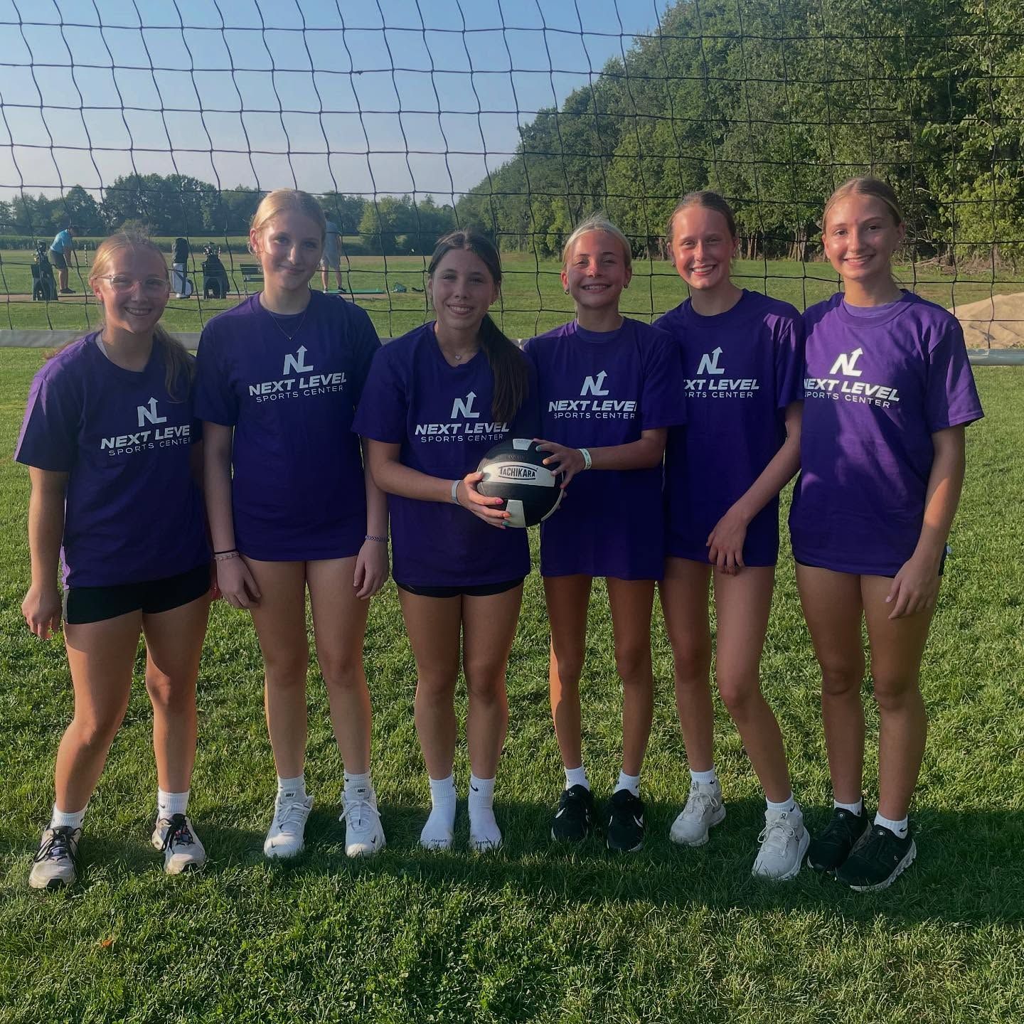 Volleyball game in an indoor dome; players in action, hitting and setting the ball. Green turf, blue ceiling.