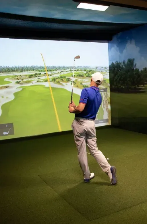 Man swinging a golf club inside a simulator, displaying a green course.