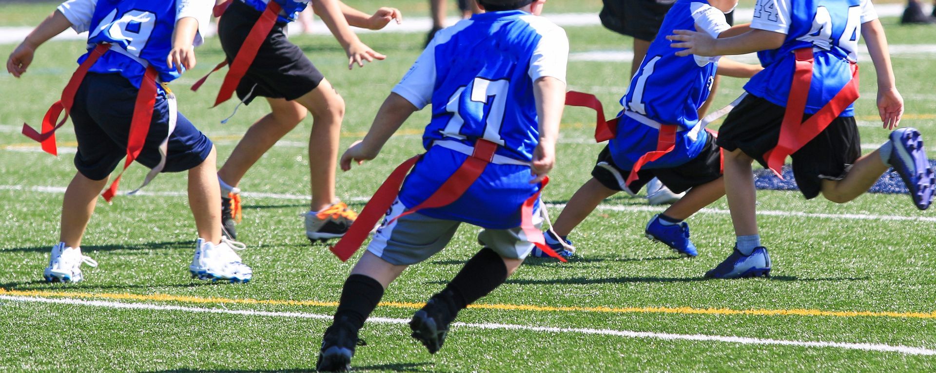 Kids in blue jerseys with red flags playing flag football on a green field.
