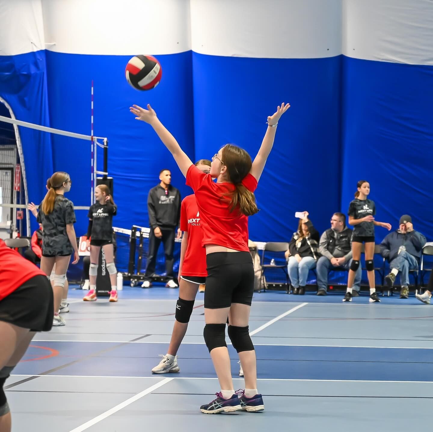 Volleyball player spikes the ball mid-air. Red and black uniforms on a red court.