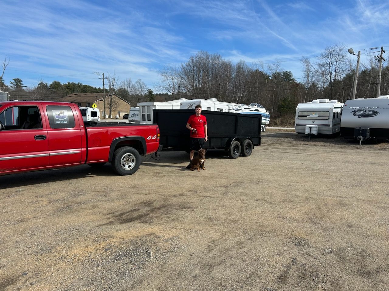 Red pickup truck towing a black trailer; a person stands nearby on a gravel lot with several RVs.