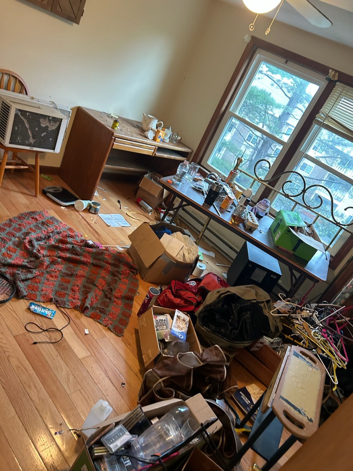 Cluttered room with wooden floors, scattered items, desk near window, blanket on floor.