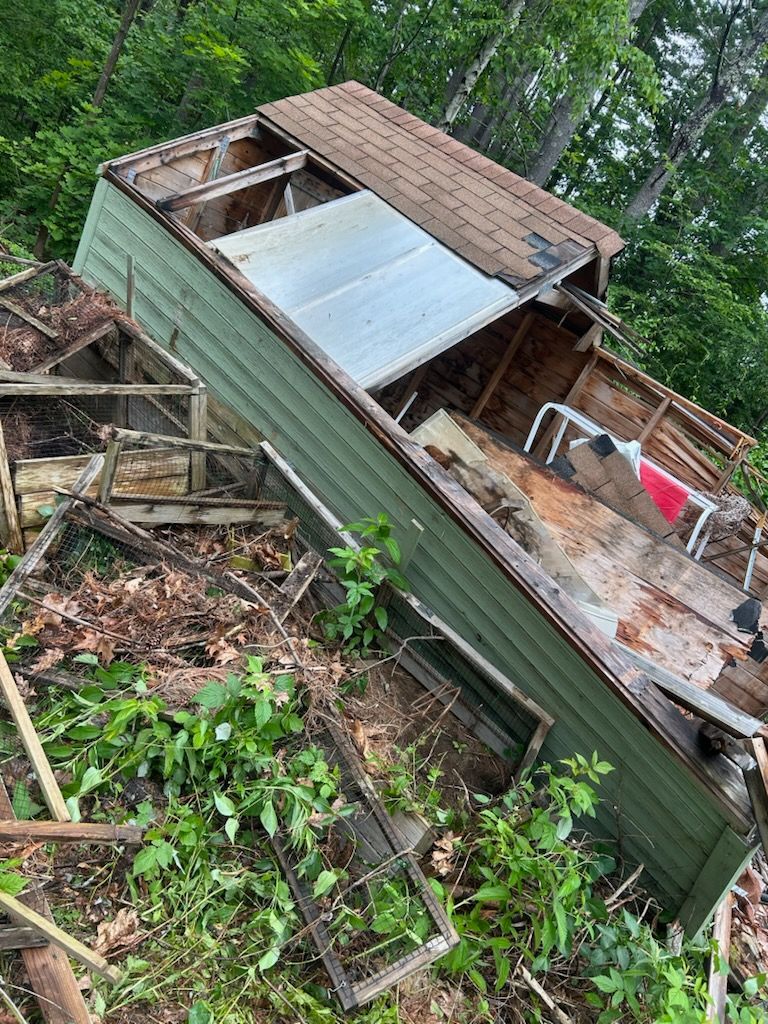 Dilapidated green shed, debris strewn on ground. Brown roof partially intact, surrounded by green foliage.