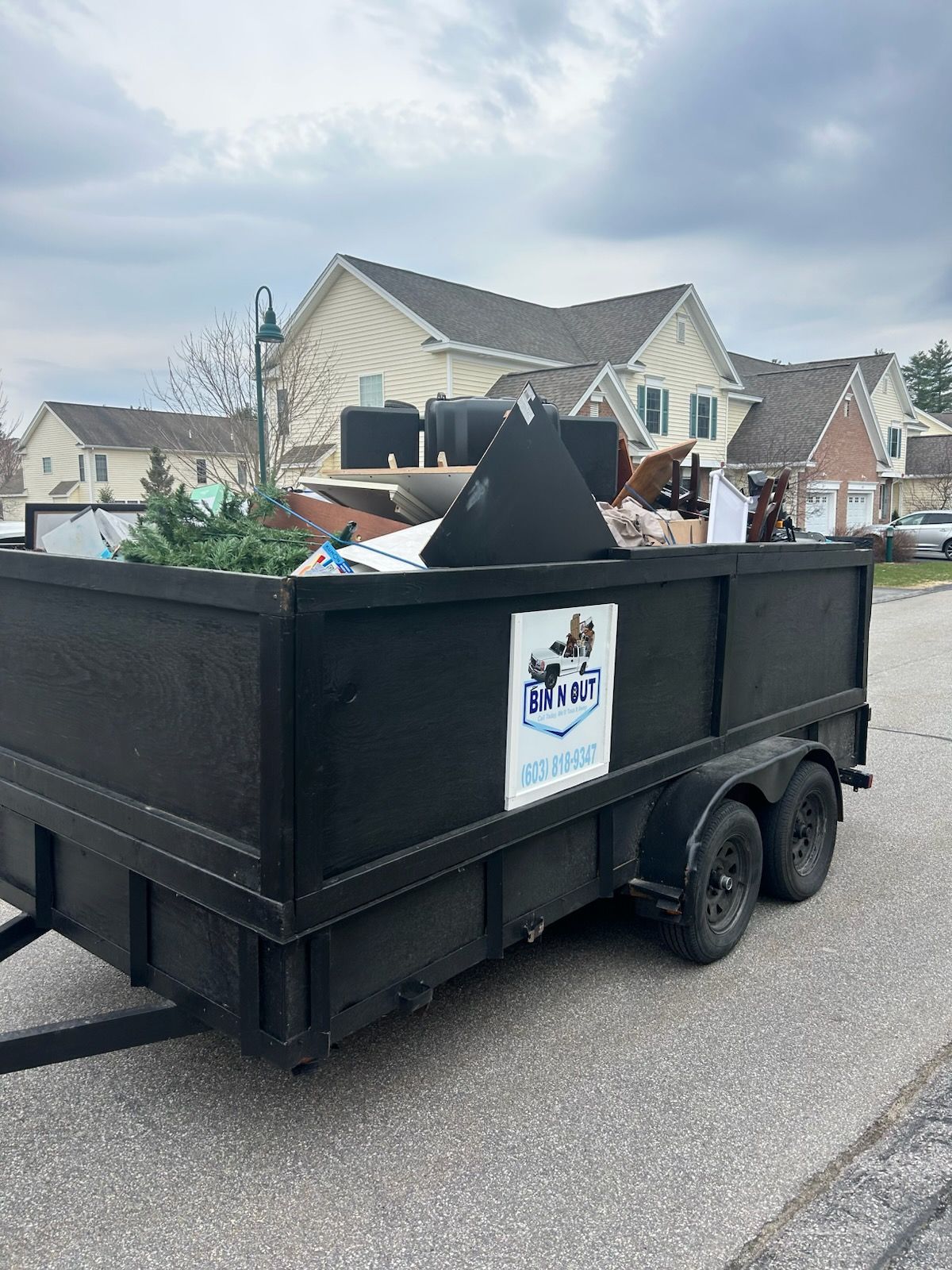 Black dumpster trailer filled with trash, parked on a street in front of houses under an overcast sky.