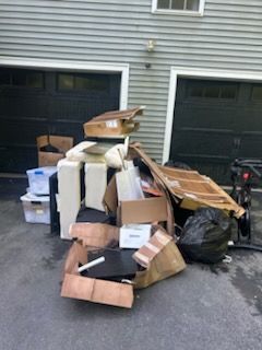 Pile of discarded boxes, furniture, and trash bags on a driveway in front of garage doors.