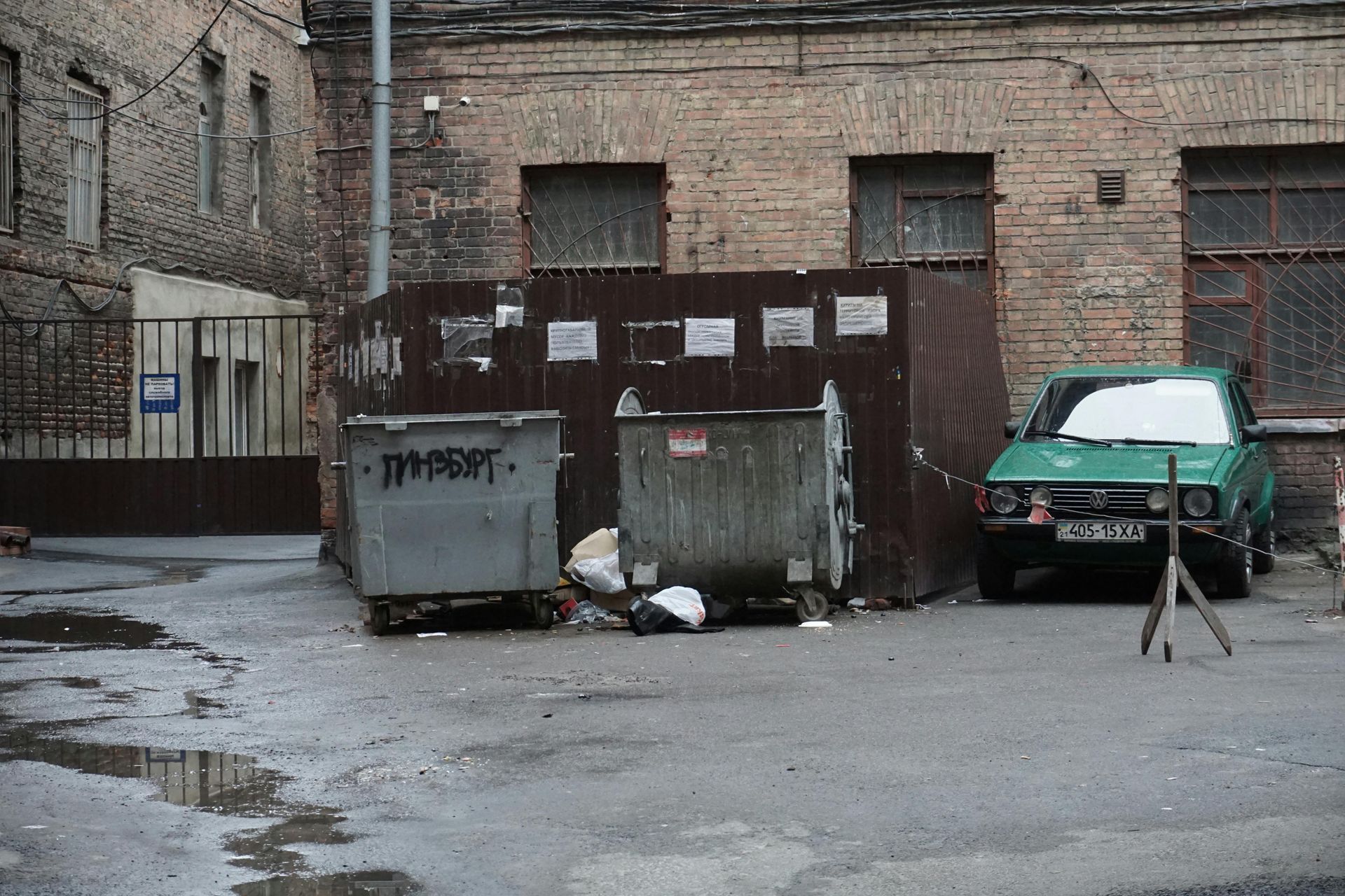 Green car parked next to dumpsters in a brick alley.