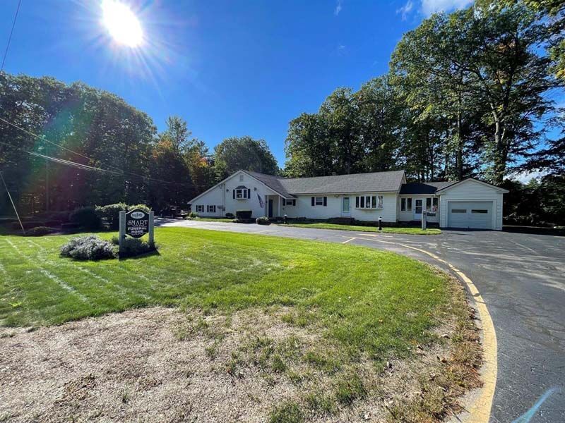 White building with garage, on a green lawn with a sign. Sunny day, blue sky, trees in the background.