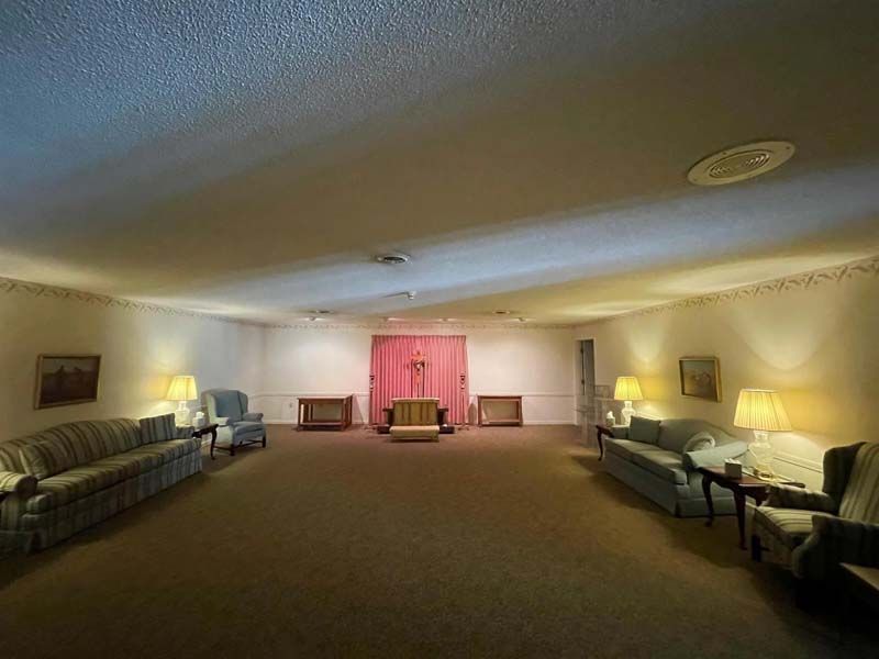 A large, beige-carpeted funeral home viewing room. Sofas and chairs face a draped altar.