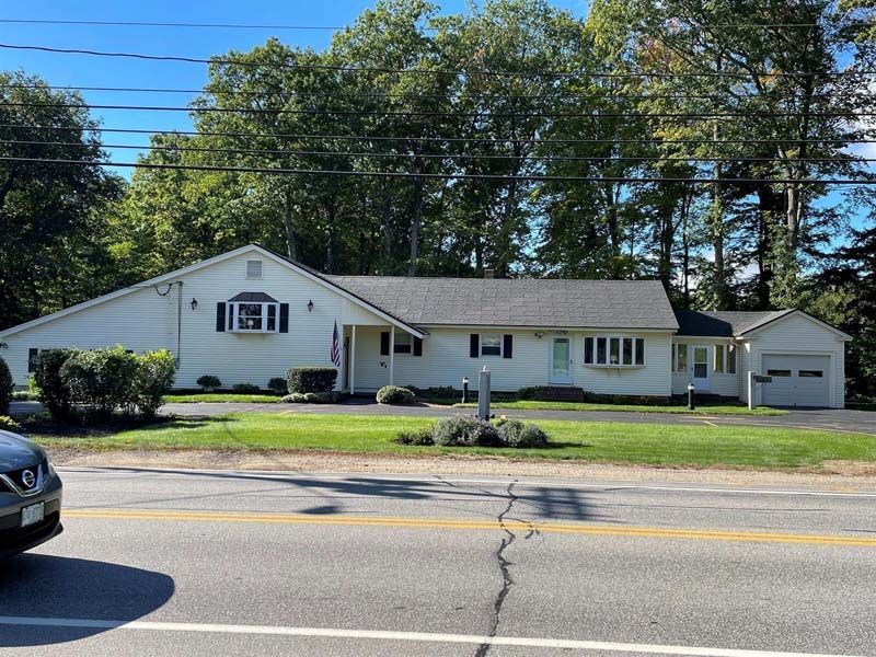 White ranch house with a garage, blue shutters, and a small lawn next to a road, trees in the background.