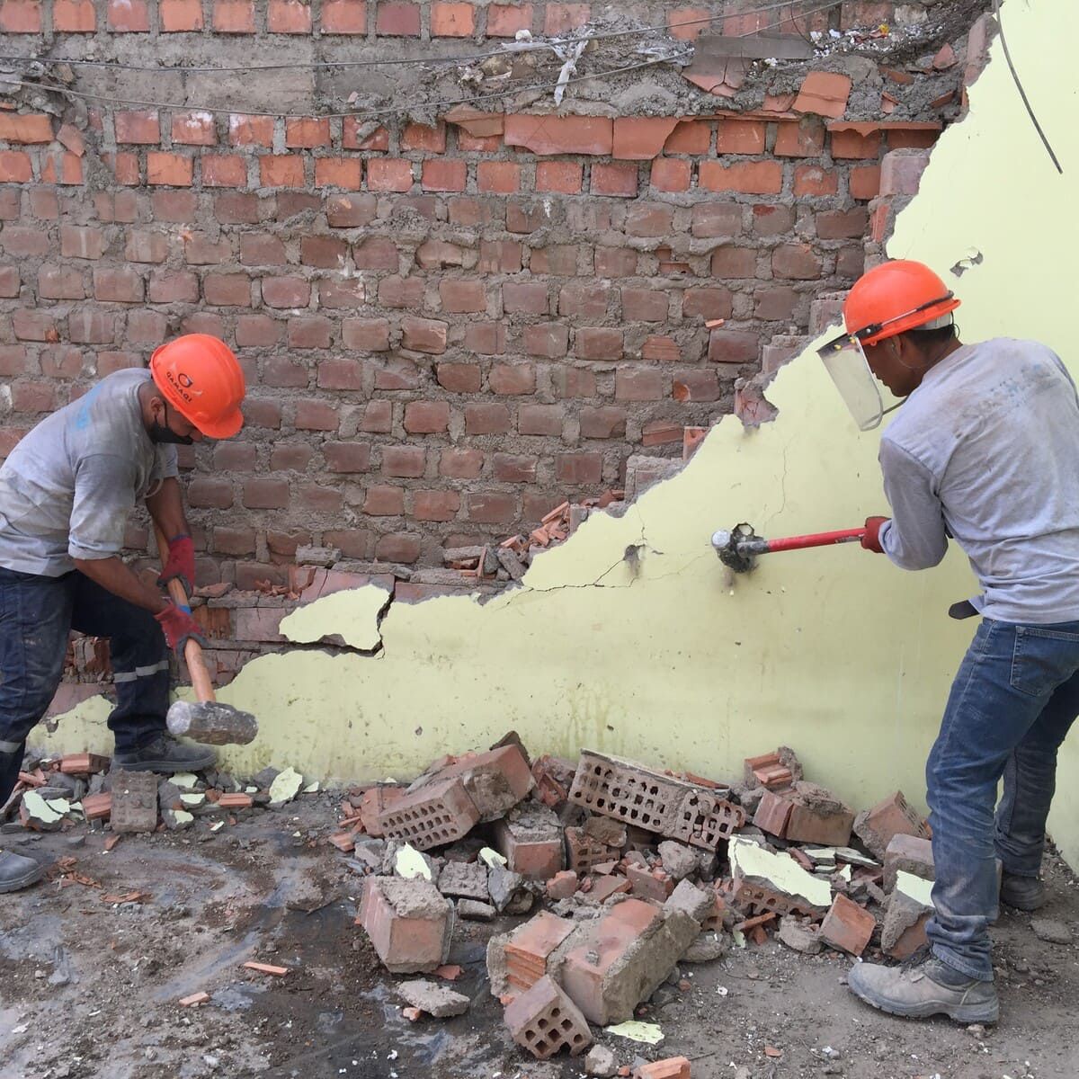 Dos trabajadores con cascos utilizan mazos para demoler una pared de ladrillos cubierta de pintura amarilla.