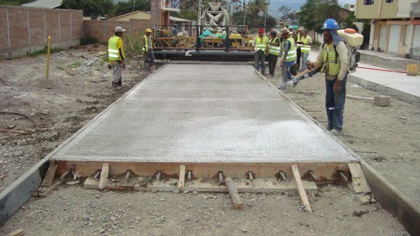 Obreros de la construcción vierten hormigón para una carretera, con equipo y un trabajador rociando un líquido.