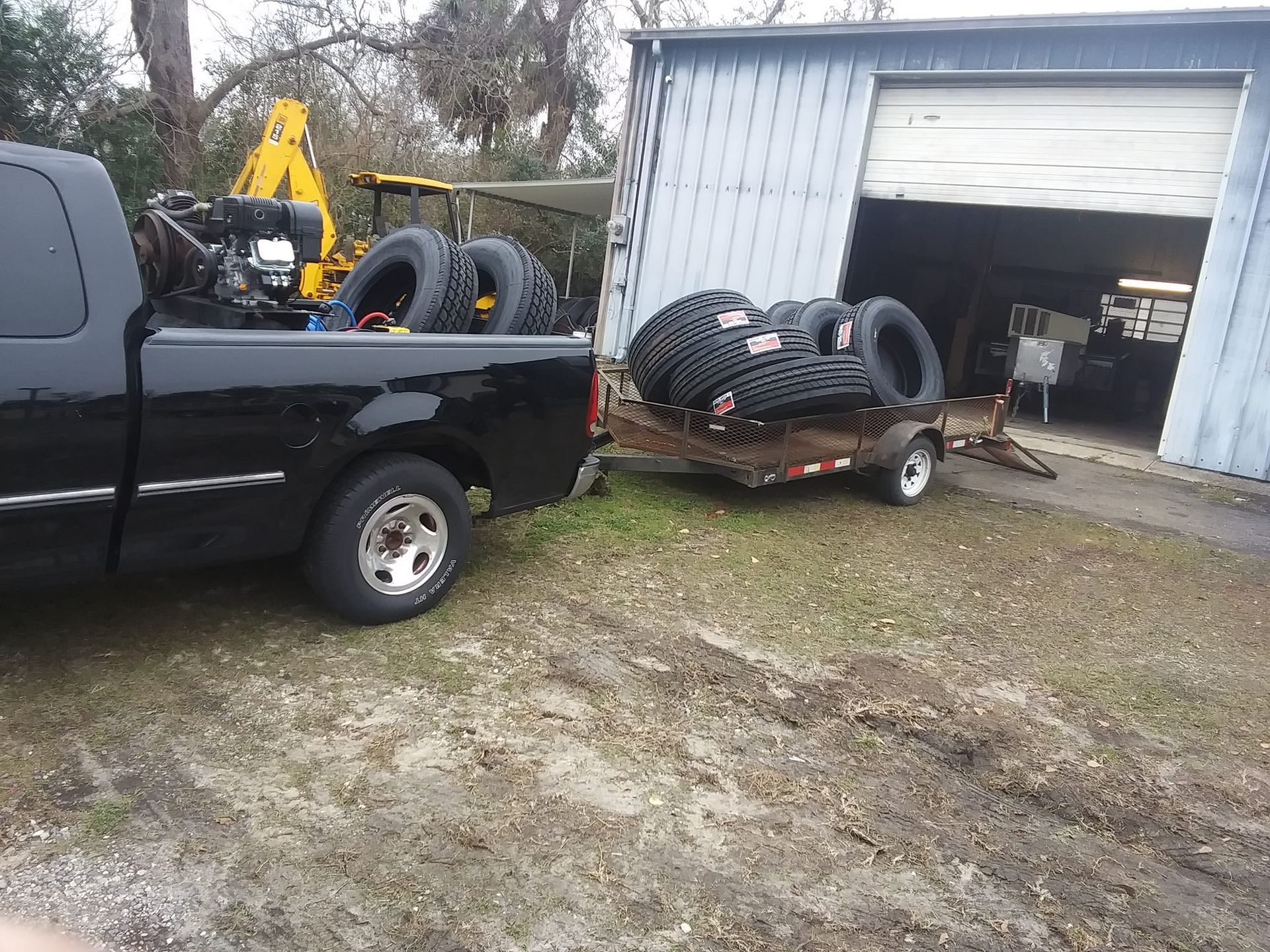 Black truck with trailer carrying tires near a garage.
