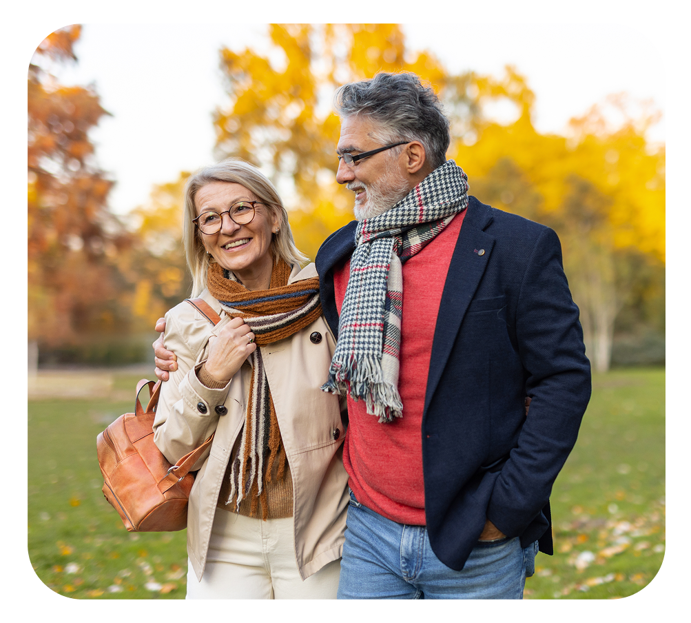Couple walking arm in arm in a park, smiling. Man in blue blazer, woman in beige coat, autumn foliage.
